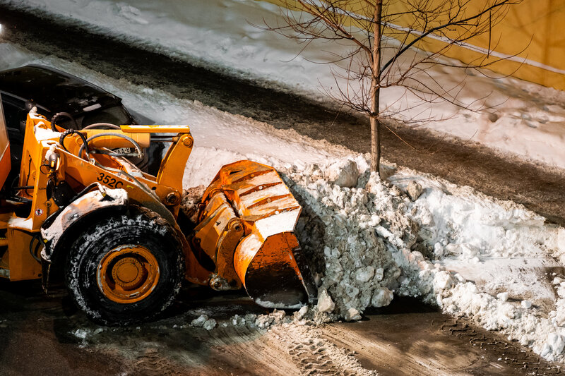 Plow truck clearing a commercial parking lot in early morning light. Heavy wet PNW snow, headlights on, cleared lanes visible.